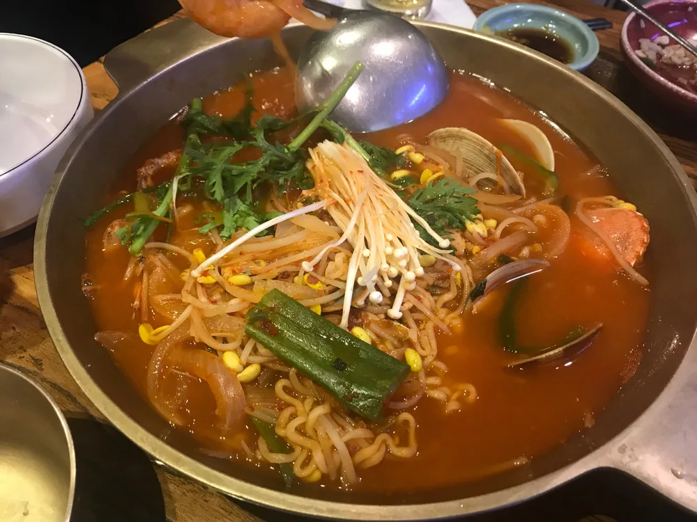 생선 라면 — 냄비에 끓인 해물라면으로, 붉은 국물에 면과 조개, 팽이버섯, 쑥갓, 콩나물, 대파가 들어 있습니다.