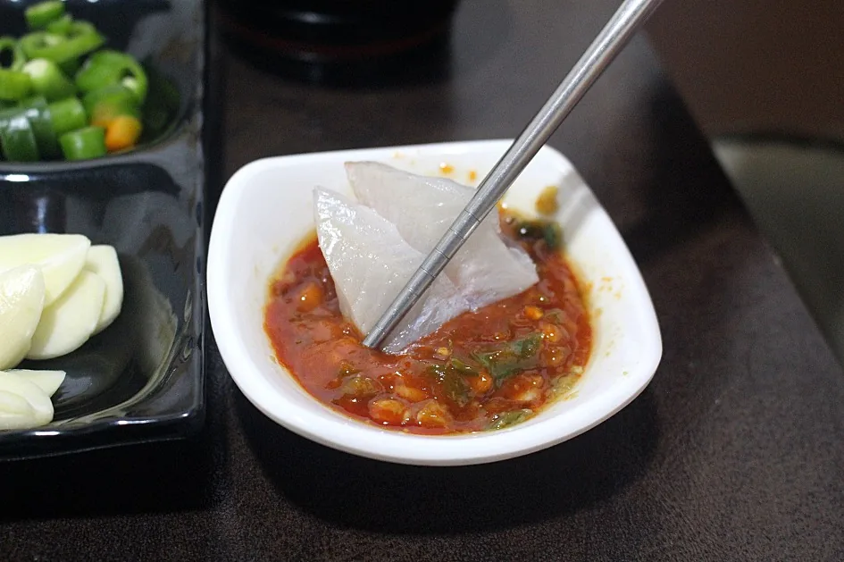 초고추장 — 볼락회 한 점을 젓가락으로 집어 초고추장과 된장을 섞은 양념에 찍고 있습니다.
